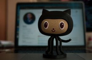 A figurine of an oktokat in the center, in the background a laptop with the main page of the GitHub open.