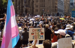 Large protest on Flinders Street in Melbourne with a trans flag and placard with the words 'Change the System' written in rainbow-coloured letters and two Aboriginal flags on it.