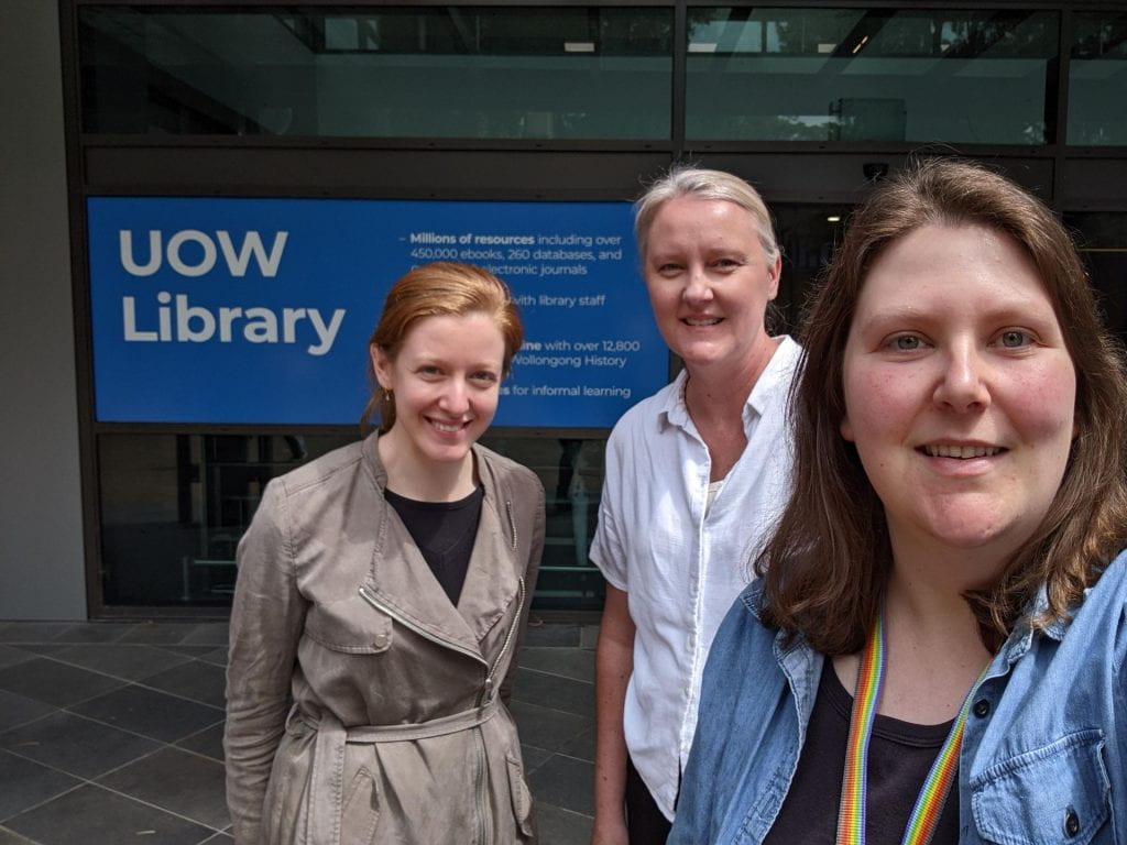 A selfie of three members of the DUX team, smiling at the camera out the front of the UOW Library building.